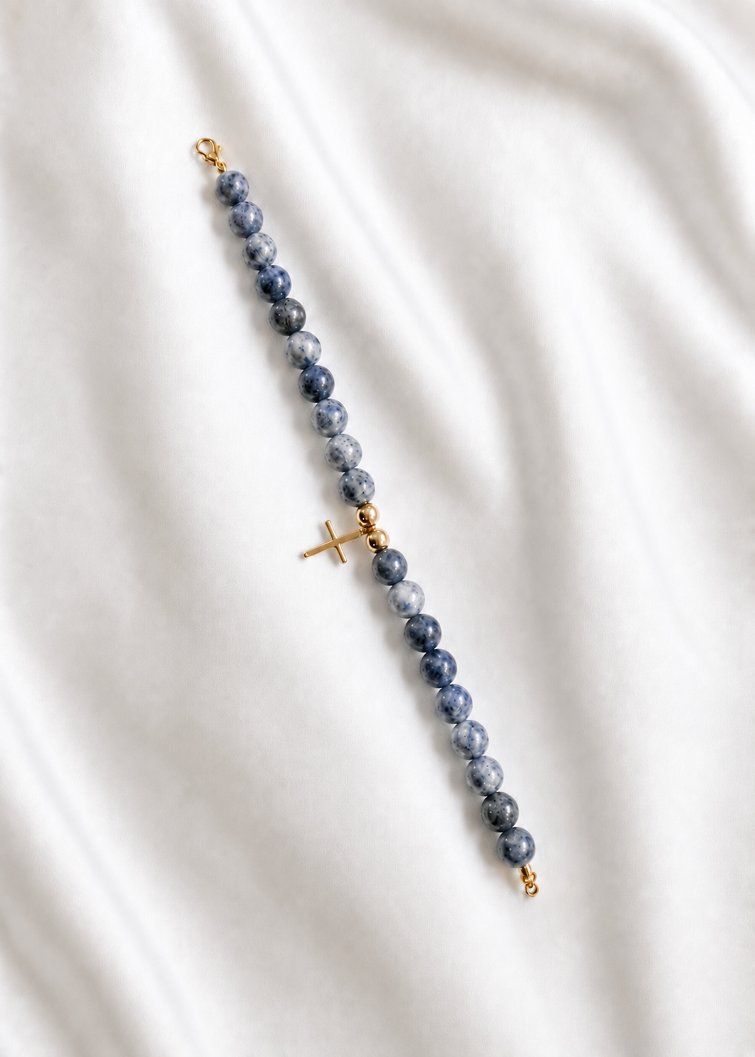 A beaded rosary-style bracelet featuring dark blue-gray sardonyx beads and gold-tone metal accents, including a central cross charm and clasps, laid diagonally on a white fabric background. Blue beaded bracelet with a gold cross, handmade and made to order