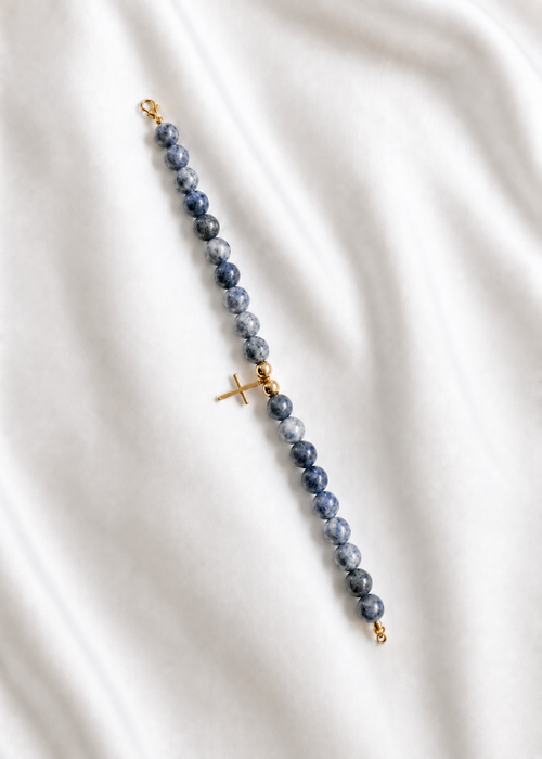A beaded rosary-style bracelet featuring dark blue-gray sardonyx beads and gold-tone metal accents, including a central cross charm and clasps, laid diagonally on a white fabric background. Blue beaded bracelet with a gold cross, handmade and made to order
