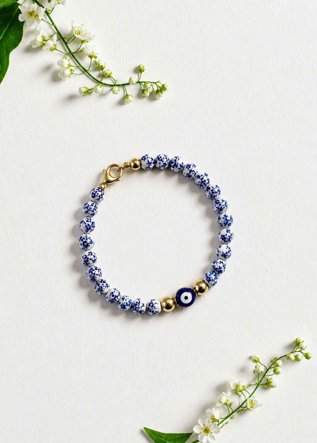 A beaded bracelet with blue and white patterned beads, a dark blue evil eye charm, and gold-tone spacer beads and clasp, arranged on a light textured background with small white flowers. Blue and white beaded bracelet featuring a blue Greek evil eye charm, inspired by Mediterranean style and the colors of Santorini. Handmade and made to order in Canada.