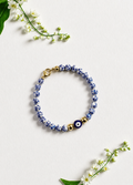 A beaded bracelet with blue and white patterned beads, a dark blue evil eye charm, and gold-tone spacer beads and clasp, arranged on a light textured background with small white flowers. Blue and white beaded bracelet featuring a blue Greek evil eye charm, inspired by Mediterranean style and the colors of Santorini. Handmade and made to order in Canada.