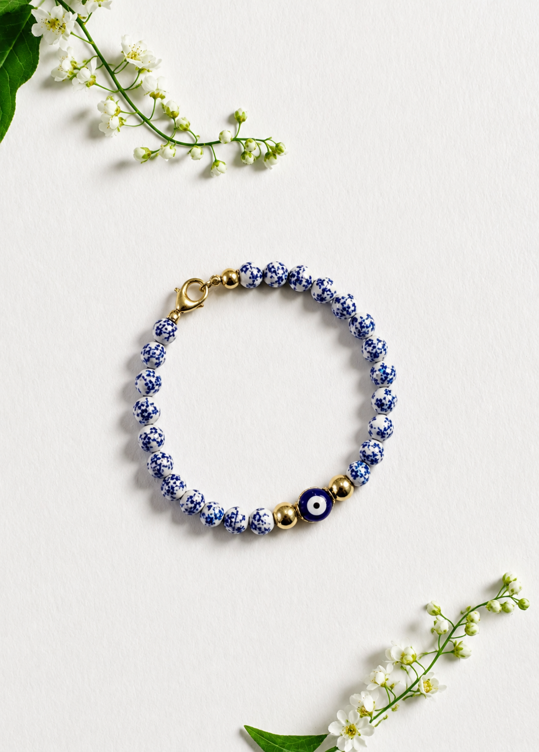 A beaded bracelet with blue and white patterned beads, a dark blue evil eye charm, and gold-tone spacer beads and clasp, arranged on a light textured background with small white flowers. Blue and white beaded bracelet featuring a blue Greek evil eye charm, inspired by Mediterranean style and the colors of Santorini. Handmade and made to order in Canada.