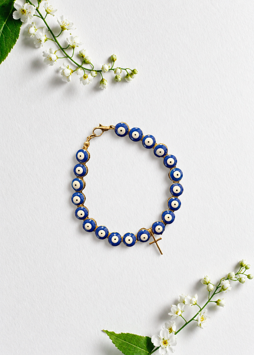A beaded evil eye bracelet with blue and white beads, gold-toned metal links, and a small gold cross charm, arranged in a flat lay on a white background with white flowers and green leaves.