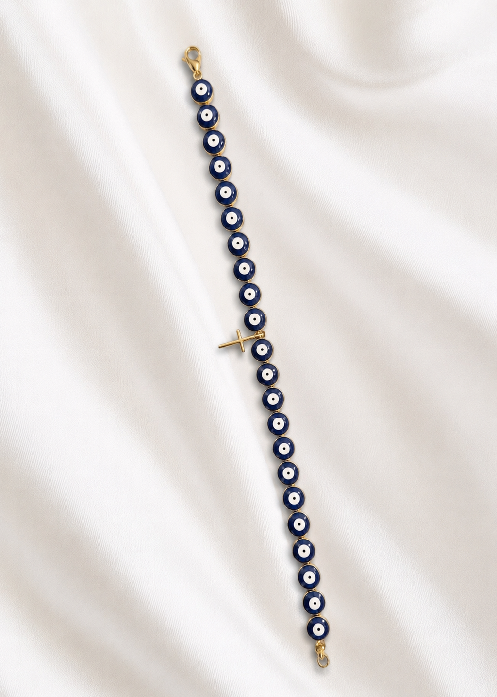 A gold-plated evil eye beaded bracelet with blue, white, and black round beads, laid out diagonally on a draped white satin fabric background.