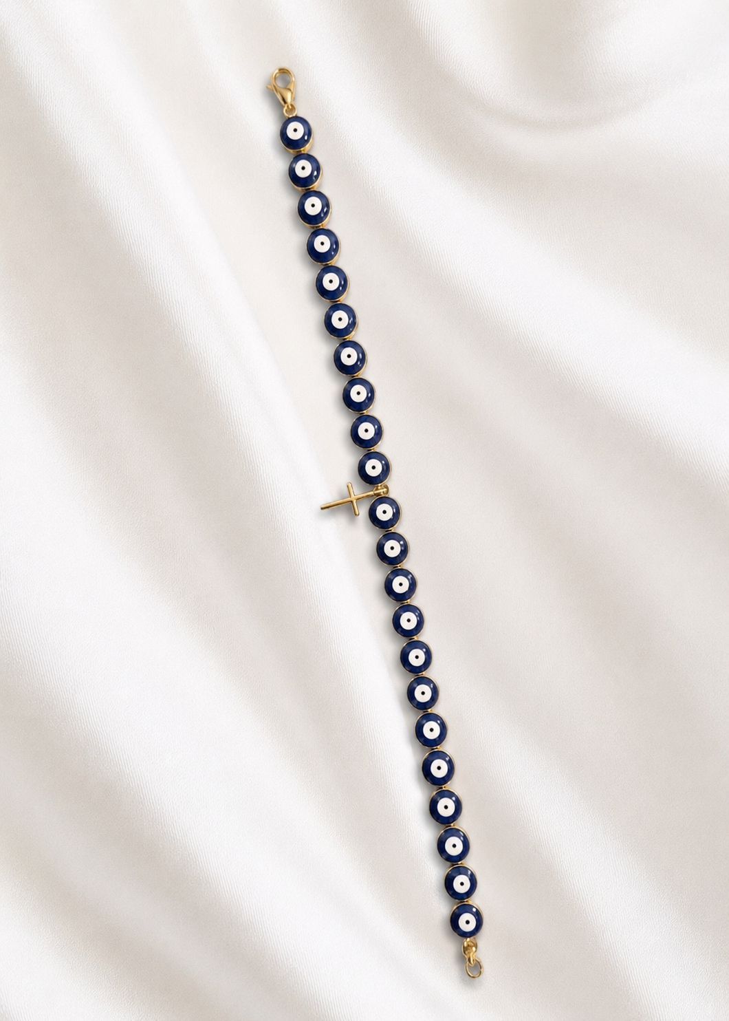 A gold-plated evil eye beaded bracelet with blue, white, and black round beads, laid out diagonally on a draped white satin fabric background.