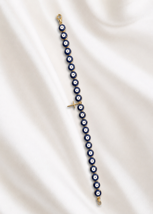 A gold-plated evil eye beaded bracelet with blue, white, and black round beads, laid out diagonally on a draped white satin fabric background.